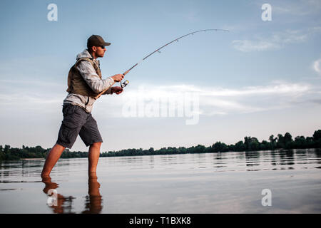 Aktive Mann steht in flachen und Angeln. Er hält die Stange in der Hand Fliegen. Der Mensch ist ein Verdrehen um die Haspel Löffel kürzer zu machen und einige Fische zu fangen. Stockfoto