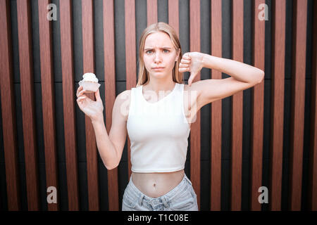 Unglückliche Mädchen steht und halten Pfannkuchen in ihrer Hand. Auch hält sie ihren großen Daumen nach unten. Auf gestreiften Hintergrund isoliert. Stockfoto