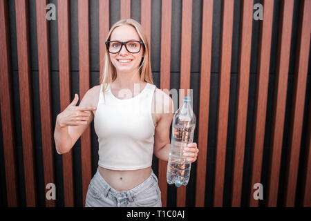 Nette und positive junge Frau steht an der Wand und Suchen auf der Kamera. Sie hält eine Flasche mit Wasser und der Große Daumen zeigen nach oben. Mädchen lächelt Stockfoto