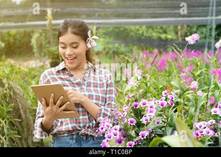 Weibliche Gärtner tragen Karohemden. Es gab Orchideen herauf die Ohren, die Hand mit der Tablette und Pointing Finger auf dem Tablett und lächelnd Stockfoto