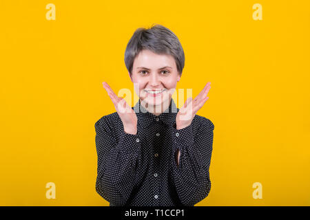 Emotionale Mädchen lächelt breit aufgeregt, breitet sich die Hände oder Applaus, im casual Hemd bekleidet, stellt gegen die gelbe Wand drückt positive Emotionen. Surpri Stockfoto