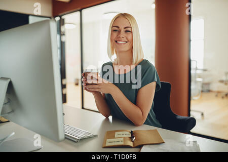 Junge Geschäftsfrau, Lächeln, während Sie an Ihrem Schreibtisch sitzen in einem Büro, trinken eine Tasse Kaffee zum Mitnehmen Stockfoto