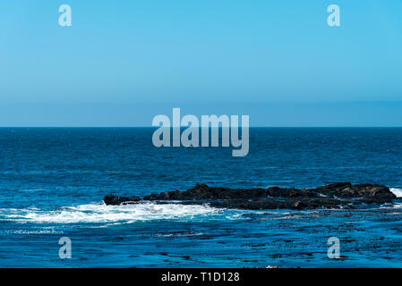 Felsige Insel im Meer mit dem Brechen der Wellen unter strahlend blauen Himmel. Stockfoto