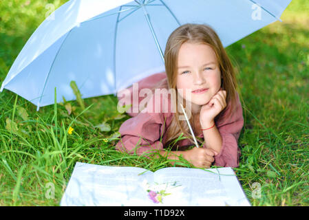 Glückliches Kind liegt auf Gras unter einem Regenschirm auf einem hellen, sonnigen Tag und liest ein Buch. Kleines Mädchen lächelnd Stockfoto