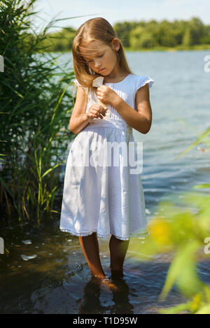 Nachdenklich, romantisch kleine Mädchen in weißem Kleid in der Nähe von Fluss hält Blume in ihren Händen. Kind steht in den Fluss Stockfoto