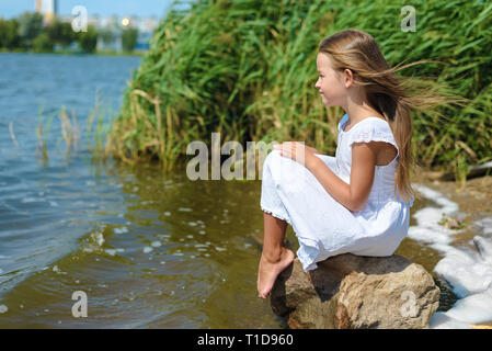 Romantisches kleines Mädchen sitzt auf Felsen in weißem Kleid an einem sonnigen Tag in der Nähe von Fluss. Das glückliche Kind ist mit der Sonne gesegnet Stockfoto