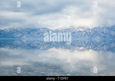 Glatte, flache Gewässer spiegeln die Kanten und die Bäume in der Ferne schneebedeckte Berge. Spiegel - Ruhig reflektierendes Wasser. Stockfoto