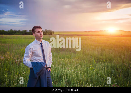Unternehmer im eleganten Anzug mit seiner Jacke hängen über seine Schulter stehend im Feld in die Ferne suchen unter einem majestätischen Abendhimmel mit einem Stockfoto
