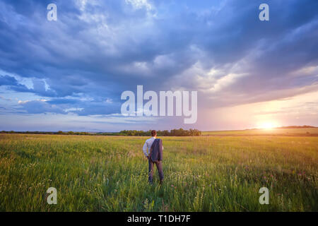 Unternehmer im eleganten Anzug mit seiner Jacke hängen über seine Schulter stehend im Feld in die Ferne suchen unter einem majestätischen Abendhimmel mit einem Stockfoto