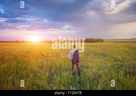 Unternehmer im eleganten Anzug mit seiner Jacke hängen über seine Schulter stehend im Feld in die Ferne suchen unter einem majestätischen Abendhimmel mit einem Stockfoto