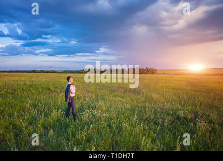 Unternehmer im eleganten Anzug mit seiner Jacke hängen über seine Schulter stehend im Feld in die Ferne suchen unter einem majestätischen Abendhimmel mit einem Stockfoto