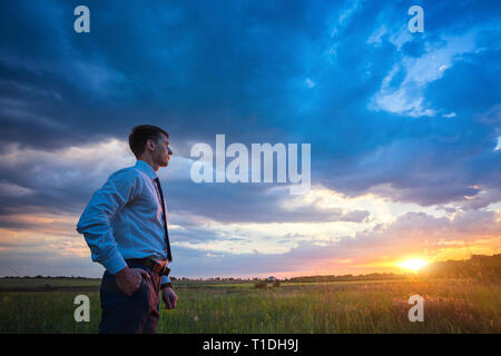 Unternehmer im eleganten Anzug mit seiner Jacke hängen über seine Schulter stehend im Feld in die Ferne suchen unter einem majestätischen Abendhimmel mit einer untergehenden Sonne. Stockfoto