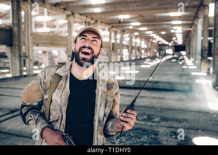 Glücklich und freundlich bärtige Kämpfer ist. Er trägt spezielle sandfarbene Uniform. Der Mensch hat, tragbares Radio in der Hand und schwarze Kerl in richtige Stockfoto