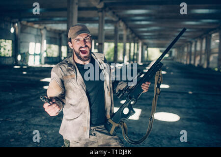 Mutig und furchtlos bärtigen Mann steht und Schreien. Er ist verrückt. Kerl hält das Gewehr in der linken Hand und Pistole in der Rechten. Mann ist verrückt. Stockfoto