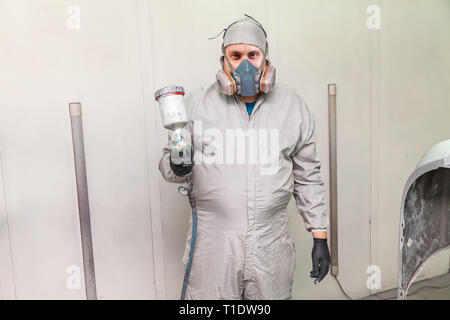 Ein männlicher Arbeitnehmer Malerei ein Auto steht in einer Spritzkabine in Schutzkleidung mit einer Spritzpistole in seine Hände nach vorne gerichtet und schauen in die c Stockfoto
