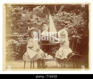 Victorian Foto der jungen Kinder im Garten tragen Hüte, (der Junge hat eine fez Stil Hut auf) spielen mit Spielzeug Yacht und modisch gekleidete Puppe in einem Kinderwagen, Großbritannien ca. 1894 Stockfoto