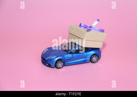 Blaue Sportwagen trägt eine Geschenkbox. Die Gabe liegt auf dem Dach des Autos. Rosa Hintergrund. Der 14. Februar. März 8. Spielzeugauto. Stockfoto
