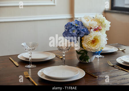 Weiße Platten, Messer und Gabeln, Kristallgläser und Blumen in Vase auf hölzernen Tisch zu Hause Stockfoto