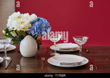 Platten, goldenen Messern und Gabeln, Kristallgläser und Blumenstrauß in weiße Vase auf hölzernen Tisch in der Nähe der roten Wand zu Hause Stockfoto