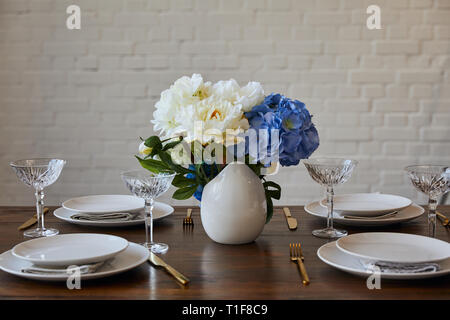 Platten, goldenen Messern und Gabeln, Kristallgläser und Blumenstrauß in weiße Vase auf hölzernen Tisch in der Nähe der Mauer zu Hause Stockfoto