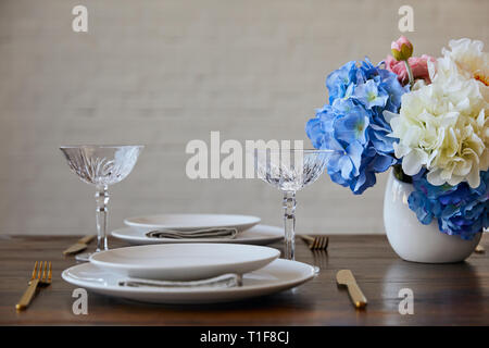 Weiße Platten, Messer und Gabeln, Kristallgläser und Blumenstrauß in Vase auf hölzernen Tisch in der Nähe der Mauer zu Hause Stockfoto