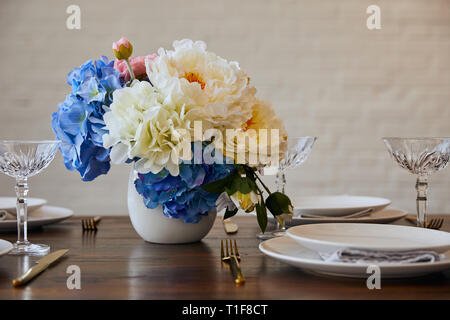 Teller, Messer und Gabeln, Kristallgläser und Blumenstrauß in weiße Vase auf hölzernen Tisch in der Nähe der Mauer zu Hause Stockfoto
