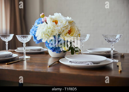 Weiß Teller, Besteck, kristall Gläser und Blumenstrauß in Vase auf hölzernen Tisch zu Hause Stockfoto