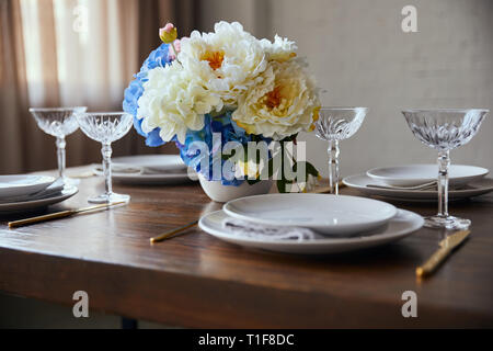 Weiße Platten, Kristallgläser und Blumen auf hölzernen Tisch zu Hause Stockfoto