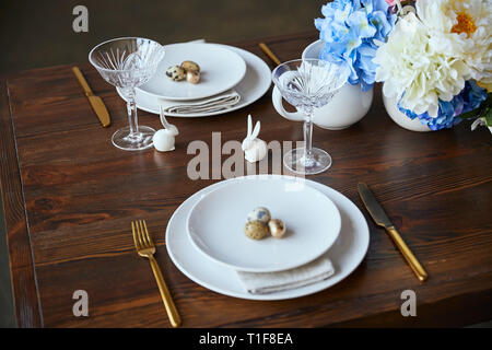 Kristallglas, Blumen in Vase, goldenes Besteck und dekorative Kaninchen auf hölzernen Tisch zu Hause Stockfoto