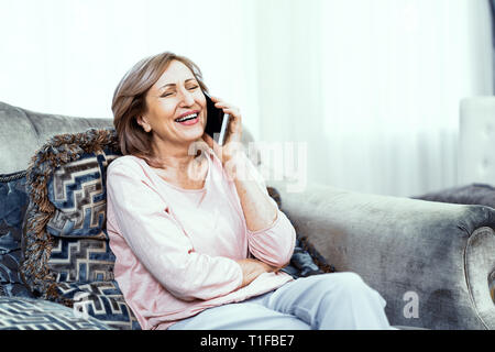 Ältere Frau mit einem Telefon in der Hand in guter Stimmung ruht sich zu Hause im Wohnzimmer. Stockfoto