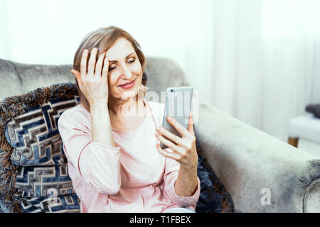 Ältere Frau mit einem Telefon in der Hand in guter Stimmung ruht sich zu Hause im Wohnzimmer. Stockfoto