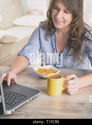 Schöne junge Frau isst Cornflakes und trinken Kaffee während der Arbeit mit dem Laptop Stockfoto