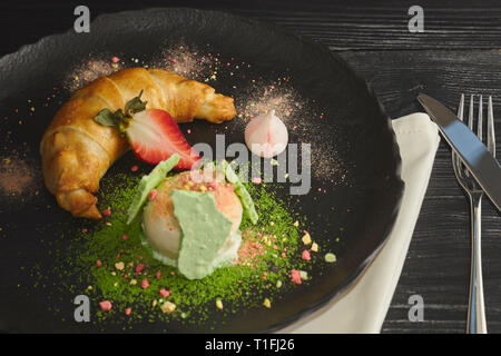 Stück Apfelstrudel mit einer Kugel Vanilleeis auf einer schwarzen Platte, auf einer hölzernen schwarz Tisch, mit Baiser und Erdbeeren dekoriert. close-up Stockfoto