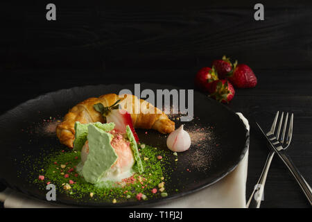 Stück Apfelstrudel mit einer Kugel Vanilleeis auf einer schwarzen Platte, auf einer hölzernen schwarz Tisch, mit Baiser und Erdbeeren dekoriert. close-up Stockfoto