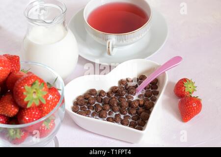 Schokolade Müsli puffs Kugeln in Milch in Herzform Schüssel als typisches Frühstück mit Saft und Erdbeeren und rosa Löffel Stockfoto