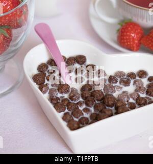 Rosa Löffel in Herzform Schüssel von Schokolade Müsli Kugeln in Milch als Detail der typischen Frühstück mit Kaffee und frische Erdbeeren Stockfoto