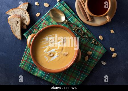 Von oben nach unten Blick auf Steingut Schüssel Kürbiscremesuppe mit Sahne auf schwarzem Hintergrund, grünen Tisch Handtuch, Messing Löffel und Tasse Früchtetee Stockfoto