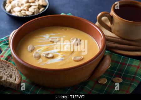 Steingut Schüssel Kürbiscremesuppe mit Sahne auf schwarzem Hintergrund, grünen Tisch Handtuch, Messing Löffel und Tasse Früchtetee Stockfoto