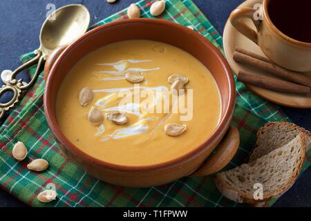 Steingut Schüssel Kürbiscremesuppe mit Sahne auf schwarzem Hintergrund, grünen Tisch Handtuch, Messing Löffel und Tasse Früchtetee Stockfoto