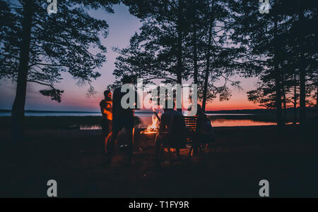 Gruppe von jungen gerne Freunde durch das Feuer im Sommer Strand sitzen, grillen Würstchen und Bier trinken, Reden und Spaß haben Stockfoto