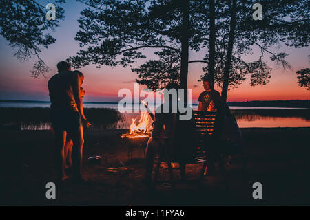 Gruppe von jungen gerne Freunde durch das Feuer im Sommer Strand sitzen, grillen Würstchen und Bier trinken, Reden und Spaß haben Stockfoto