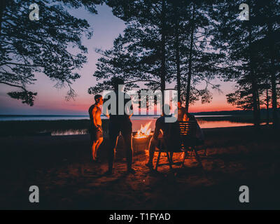 Gruppe von jungen gerne Freunde durch das Feuer im Sommer Strand sitzen, grillen Würstchen und Bier trinken, Reden und Spaß haben Stockfoto