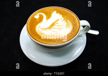 Cappuccino mit Schwan auf dem Schaumstoff gezogen, in eine weiße Tasse mit Löffel und Untertasse, schwarzer Hintergrund Stockfoto