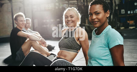 Zwei passen junge Frauen in Sportkleidung Lächeln beim Sitzen mit ein paar Freunde auf dem Boden einer Turnhalle nach dem Training Stockfoto