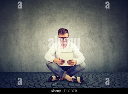 Lächelnden jungen Mann in Gläser mit einem Tablet sitzen auf einem Stock entspannt und in einer Videokonferenz über graue Wand Hintergrund Stockfoto
