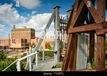 Mit Blick auf die Innenstadt von einer Mine Fördergerüst über eine alte Mine Welle mit einem historischen und Gedenk- Display, in Butte, MT, USA Stockfoto