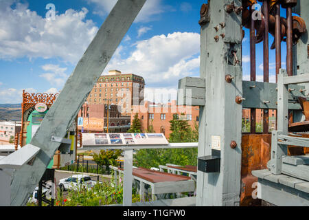 Mit Blick auf die Innenstadt von einer Mine Fördergerüst über eine alte Mine Welle mit einem historischen und Gedenk- Display, in Butte, MT, USA Stockfoto