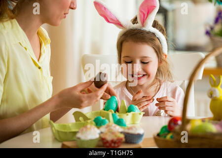 Mutter und Tochter Ostern feiern, Schokolade essen Eier. Happy Family Urlaub. Süße kleine Mädchen mit lustigen Gesicht in Hasenohren lachen, lächeln Stockfoto