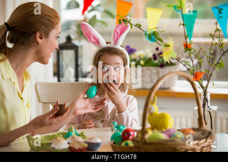 Mutter und Tochter Ostern feiern, Schokolade essen Eier. Happy Family Urlaub. Süße kleine Mädchen mit lustigen Gesicht in Hasenohren lachen, lächeln Stockfoto