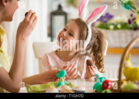 Mutter und Tochter Ostern feiern, Schokolade essen Eier. Happy Family Urlaub. Süße kleine Mädchen mit lustigen Gesicht in Hasenohren lachen, lächeln Stockfoto